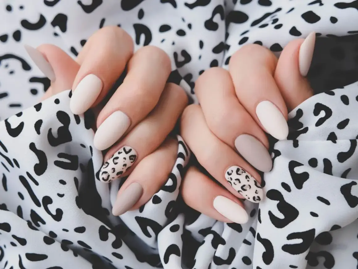 Dłonie z eleganckim panterkowym manicure w odcieniach beżu i ecru na tle wzorzystej tkaniny.