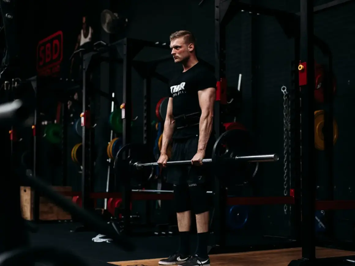 Ein Mann mit Bart und schwarzem T-Shirt hebt eine Langhantel im Fitnessstudio. Er trainiert seine Handkraft, um stärker zu werden.