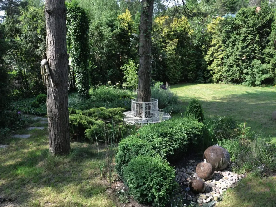 Urokliwy ogród w lesie z kamiennymi kulami tworzącymi wodną kaskadę, ozdobną ławką wokół drzewa i bujną zielenią.