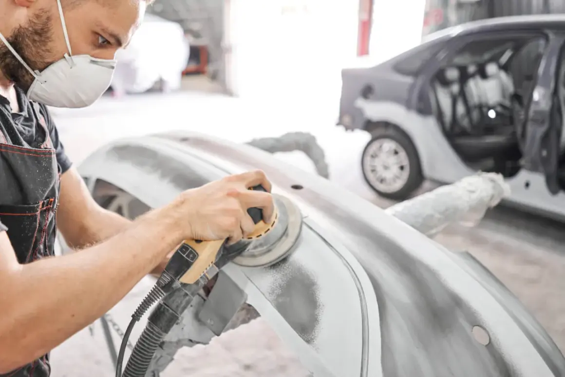 Pracownik w masce ochronnej wykonuje szlifowanie szpachli samochodowej na zderzaku. W tle widać auto w trakcie naprawy.