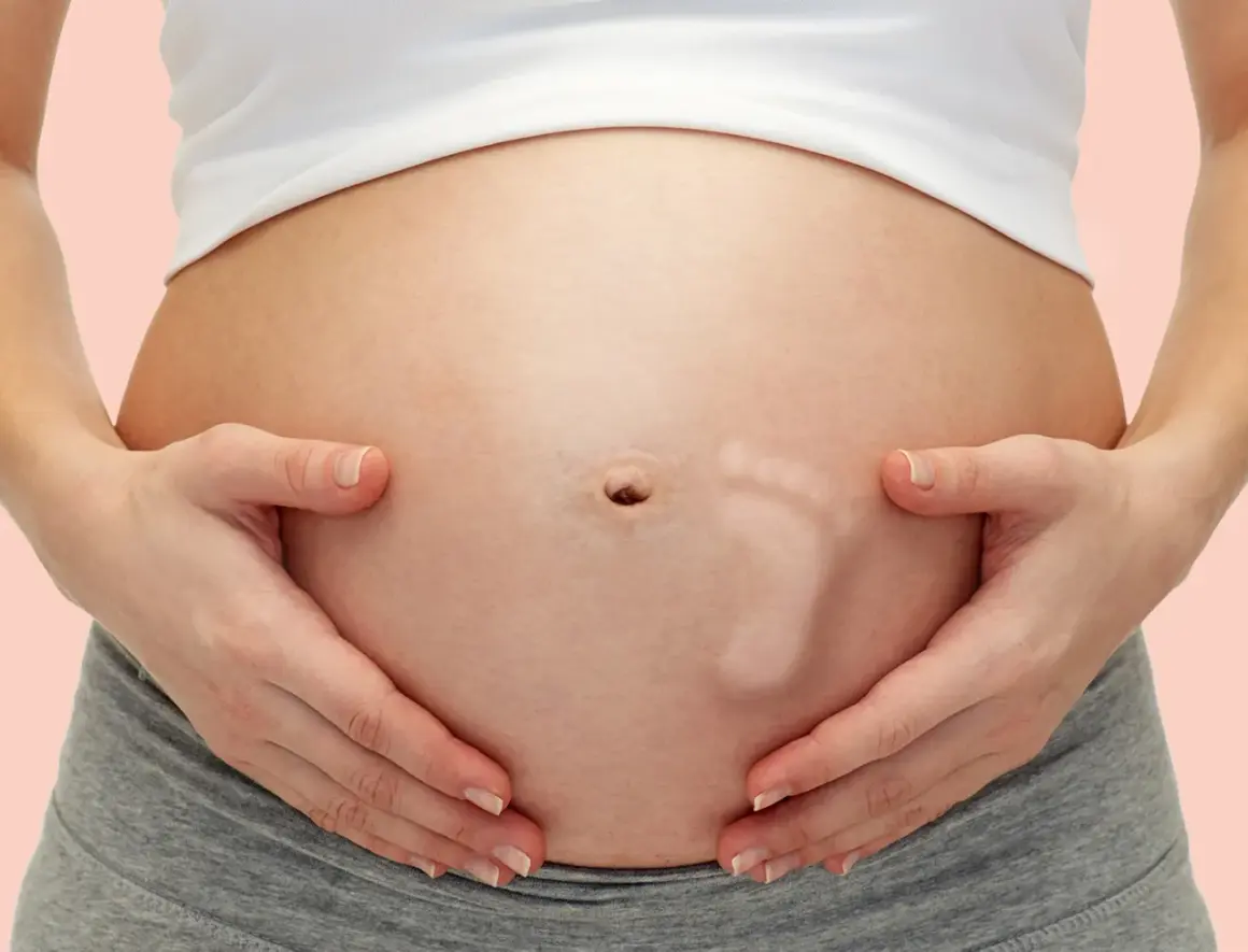 Brzuszek w 29 tygodniu ciąży, z widocznym odciskiem stópki dziecka. Ręce delikatnie obejmują brzuch.