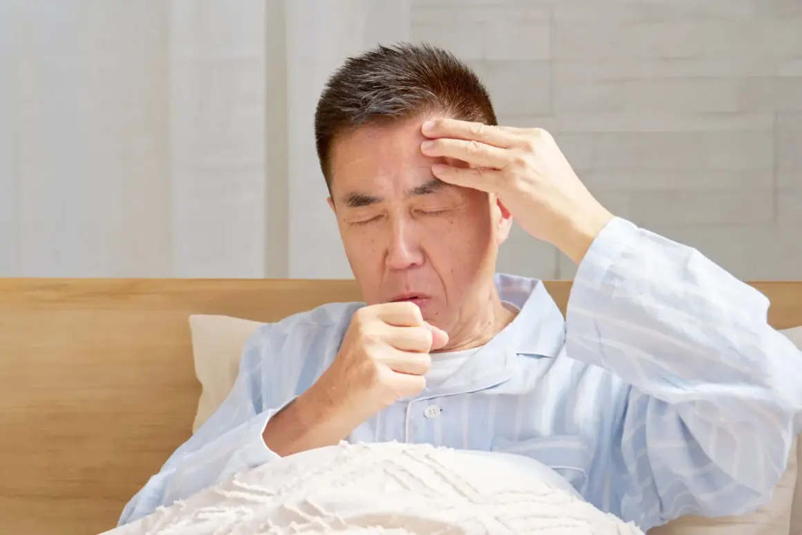 Mann im Bett mit geschlossenen Augen, der sich die Stirn reibt und hustet. Er leidet unter Kopfschmerzen beim Husten.
