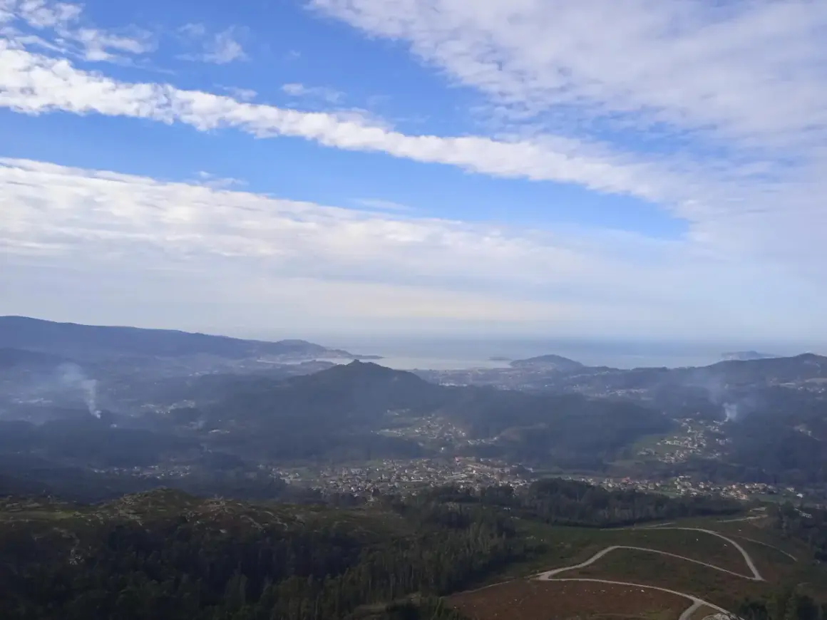 Vista panorámica desde el monte Galiñeiro, con un valle salpicado de casas y el mar al fondo bajo un cielo nublado.