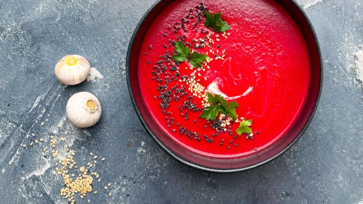 Eine leuchtend rote Bete Suppe, garniert mit Sesam und Petersilie, neben zwei Knoblauchzehen.