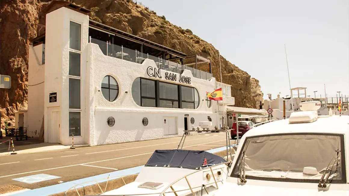 Edificio blanco con el letrero "C.N. SAN JOSE" y una bandera española ondeando. Un restaurante con vistas al mar, ideal para disfrutar de la brisa marina.