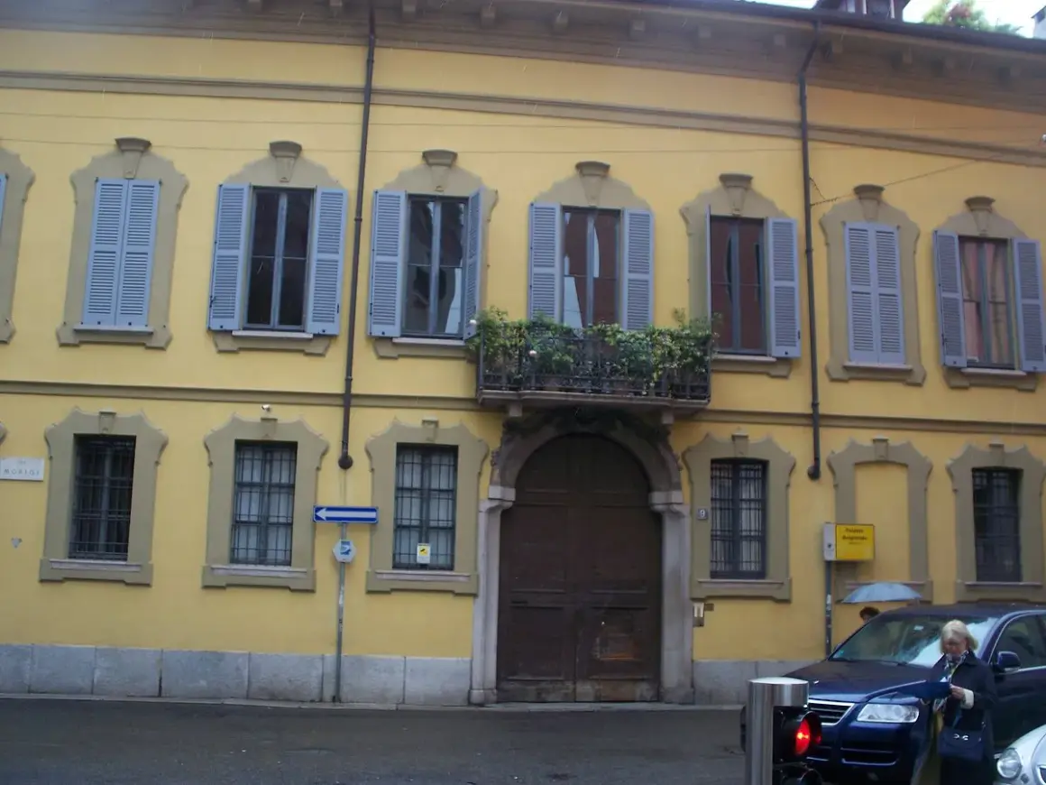 Facciata gialla del **Palazzo Belgioioso** con finestre dalle persiane azzurre e un balcone fiorito.