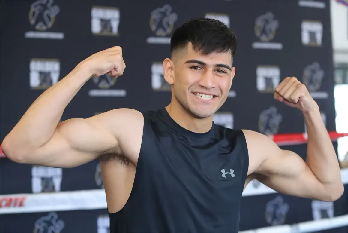Alexis boxer flexes his biceps, smiling confidently in a black tank top.
