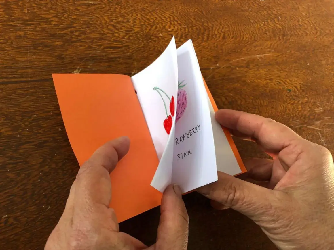 Mani che sfogliano un piccolo libro fatto a mano, con disegni di fragole e ciliegie. Un'idea per imparare come fare un libro a mano.