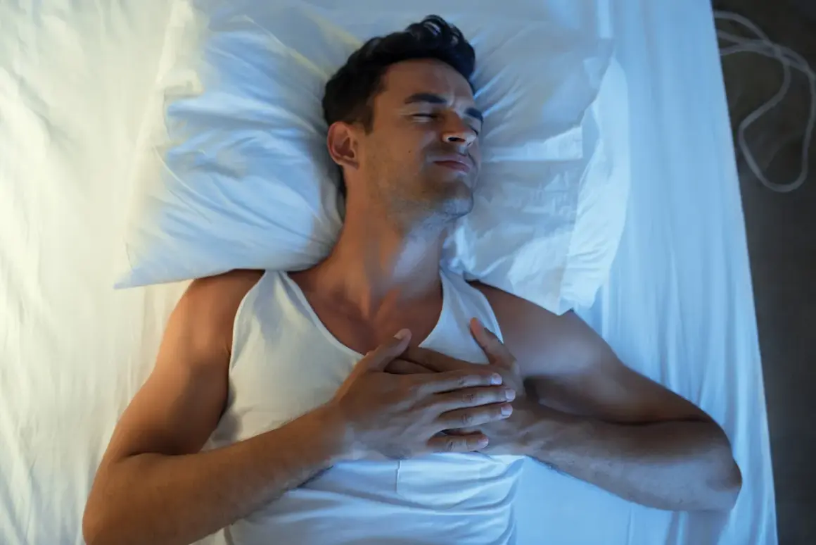 Mann liegt im Bett, hält sich die Brust. Er leidet unter Herzrasen nachts, die Augen sind geschlossen, das Gesicht verzogen.