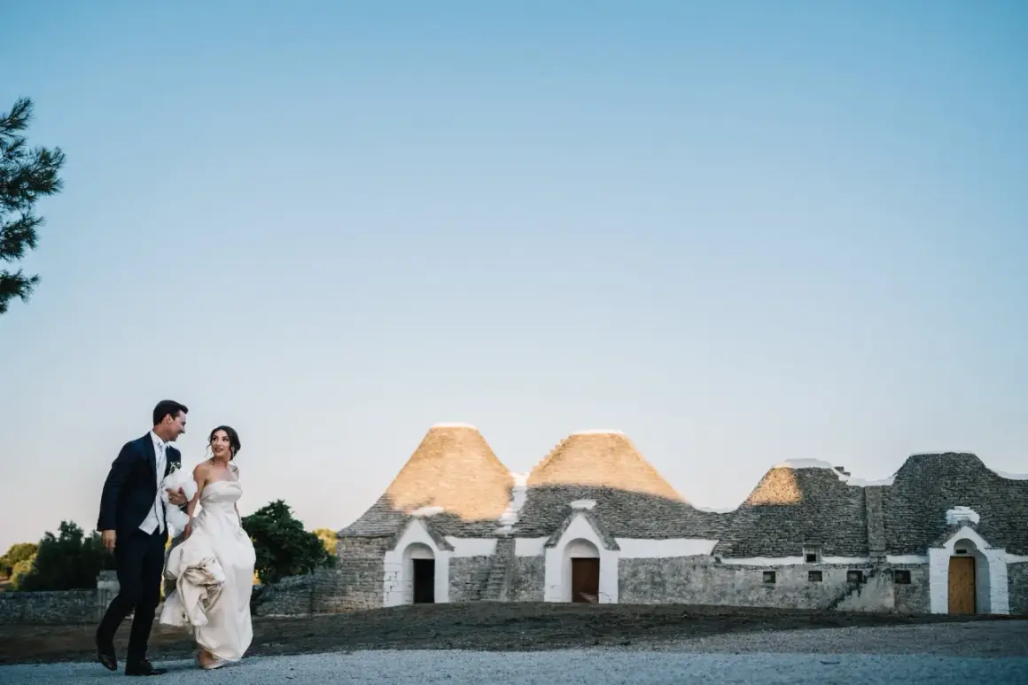 Sposi passeggiano sorridenti davanti a trulli, un'immagine perfetta per le canzoni ingresso sposi.