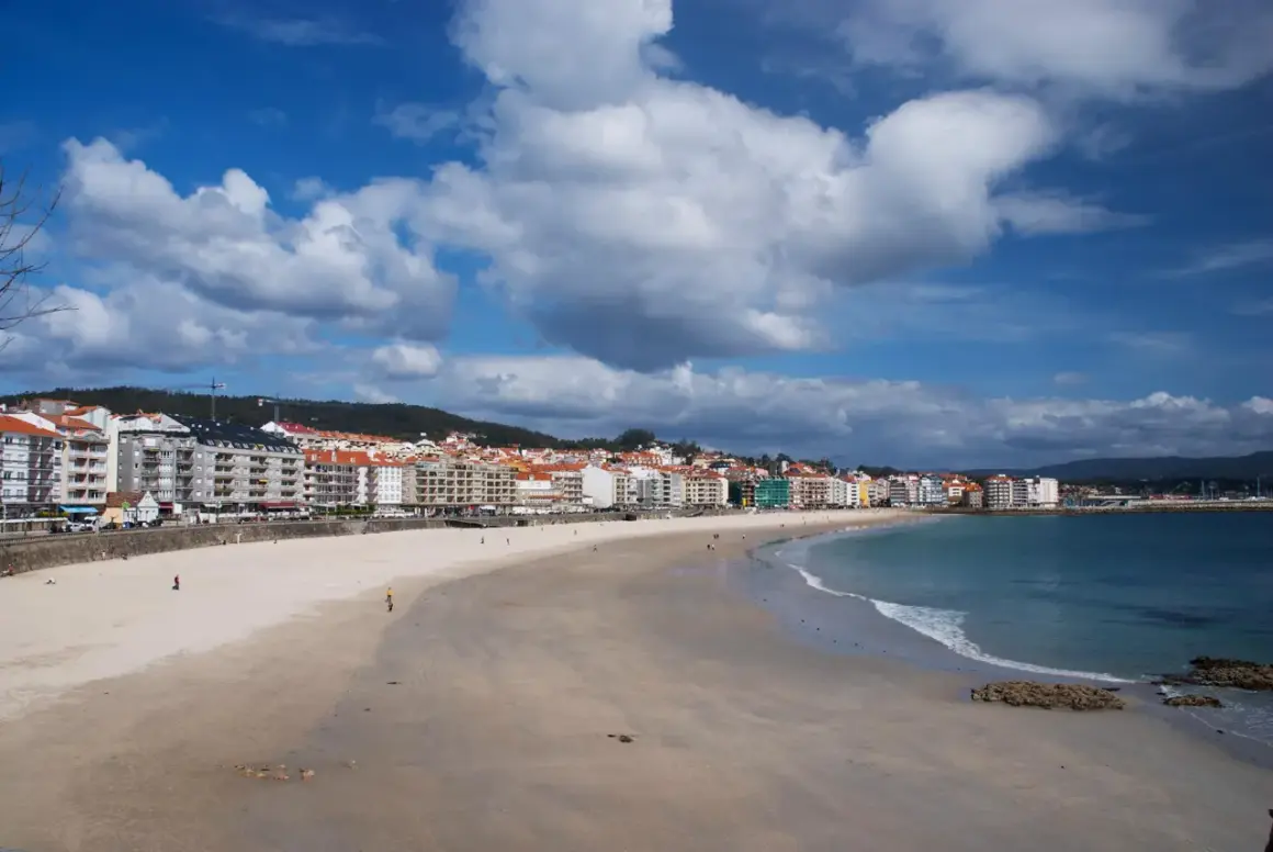Amplia playa de Silgar con edificios residenciales al fondo y el mar azul bajo un cielo nublado.