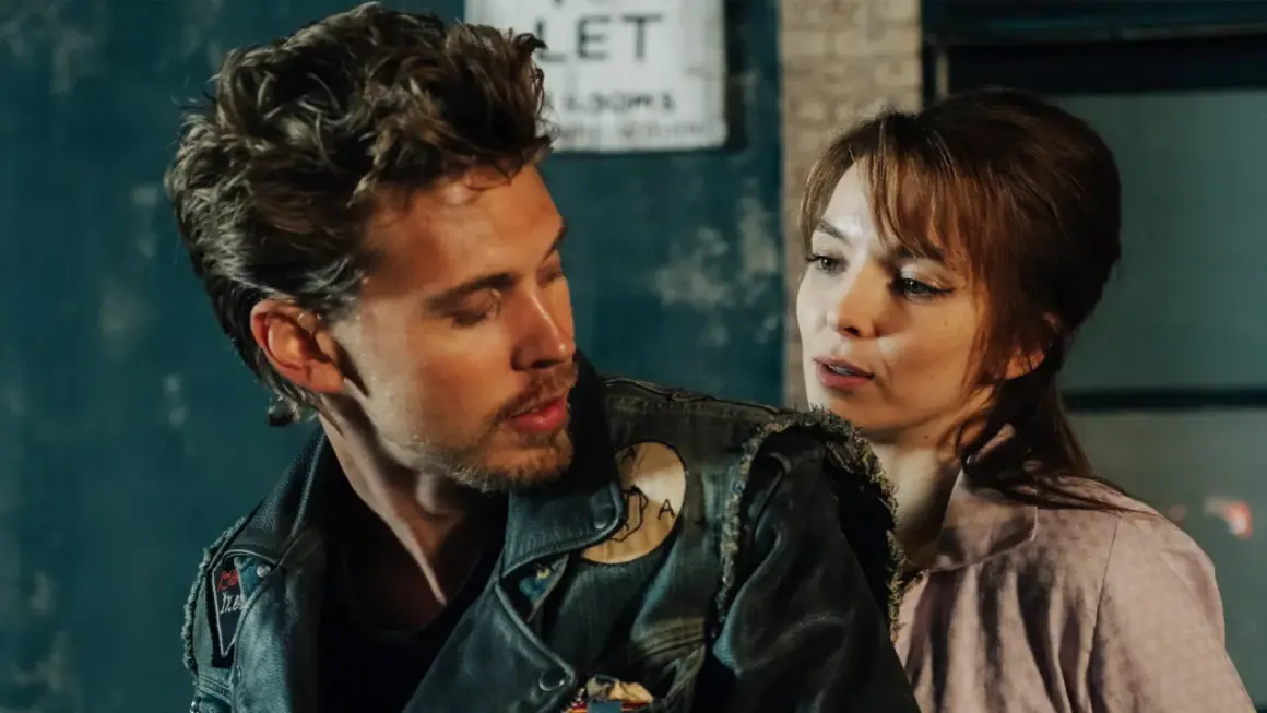Ein Mann mit Lederjacke und eine Frau im rosa Hemd blicken sich an. Szene aus "The Bikeriders" Stream.
