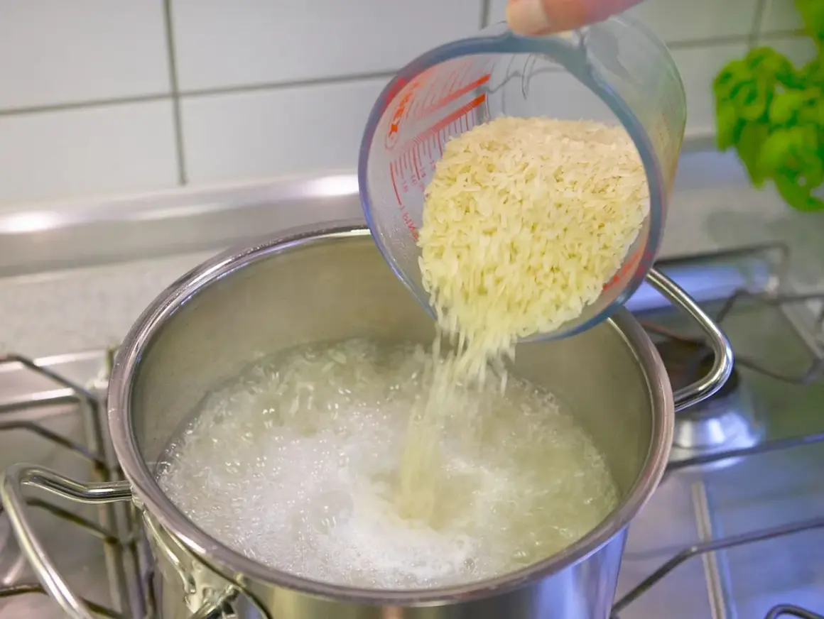 Reis im Topf kochen: Eine Hand schüttet Reis aus einem Messbecher in kochendes Wasser.