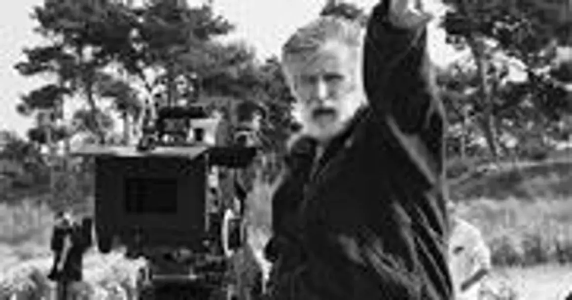 Edward Kłosiński, z siwą brodą, gestykuluje na planie filmowym. W tle widać kamerę i drzewa.