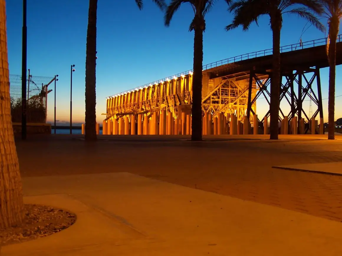 Atardecer en el parque de las almadrabillas, con palmeras y un puente iluminado sobre el mar.