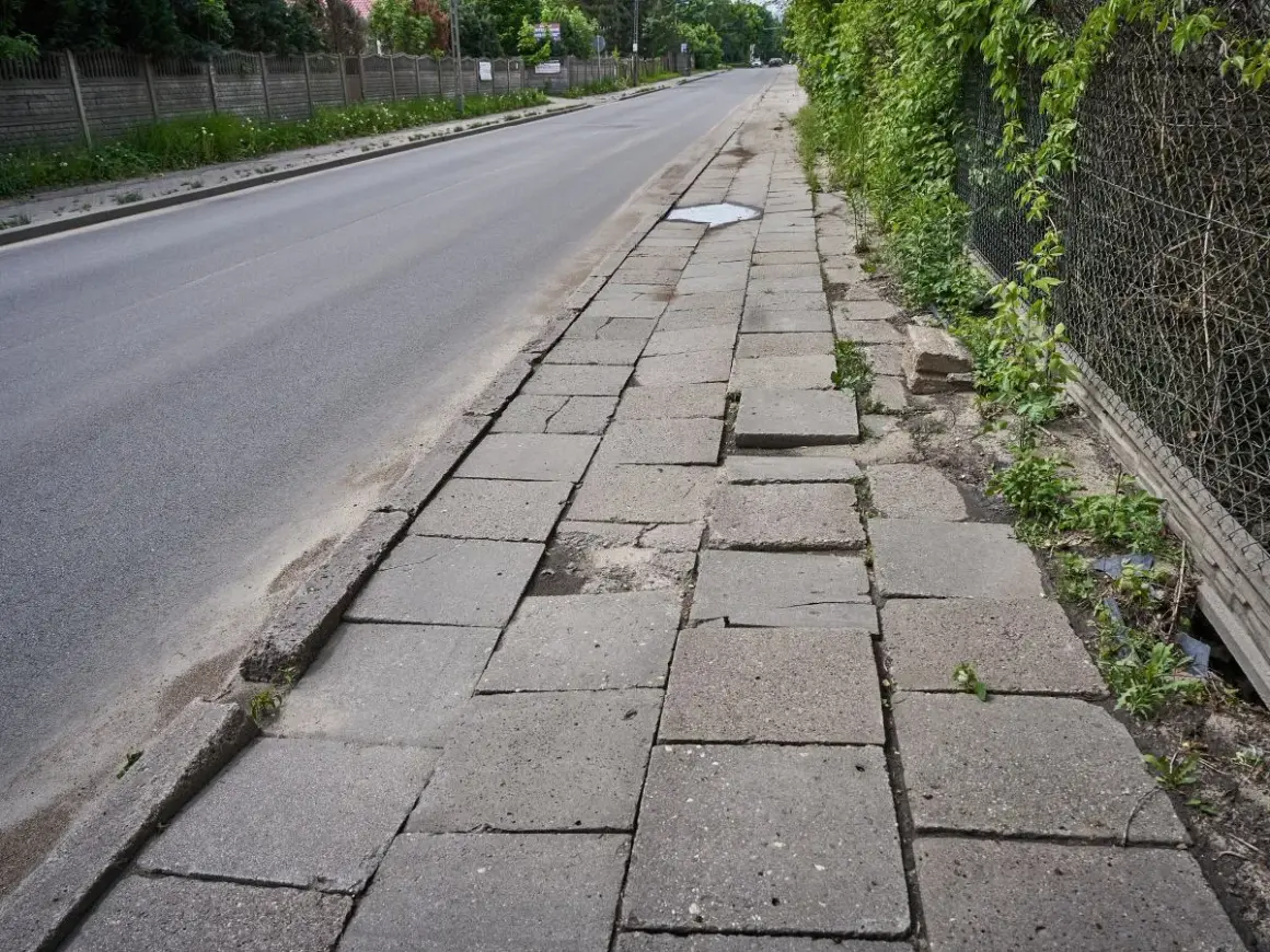 Zniszczony chodnik obok drogi. Płyty chodnikowe są popękane i nierówne, tworząc niebezpieczną ścieżkę dla pieszych.