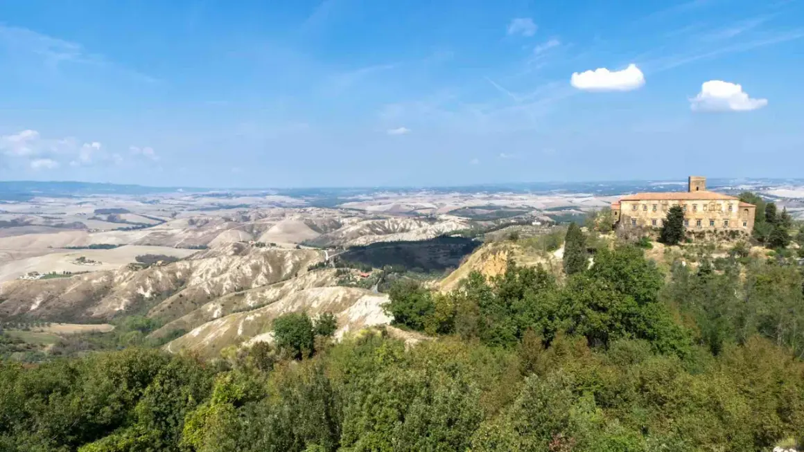 Panoramica delle **balze di Volterra**, con un casale in pietra in primo piano e dolci colline ondulate all'orizzonte sotto un cielo azzurro.