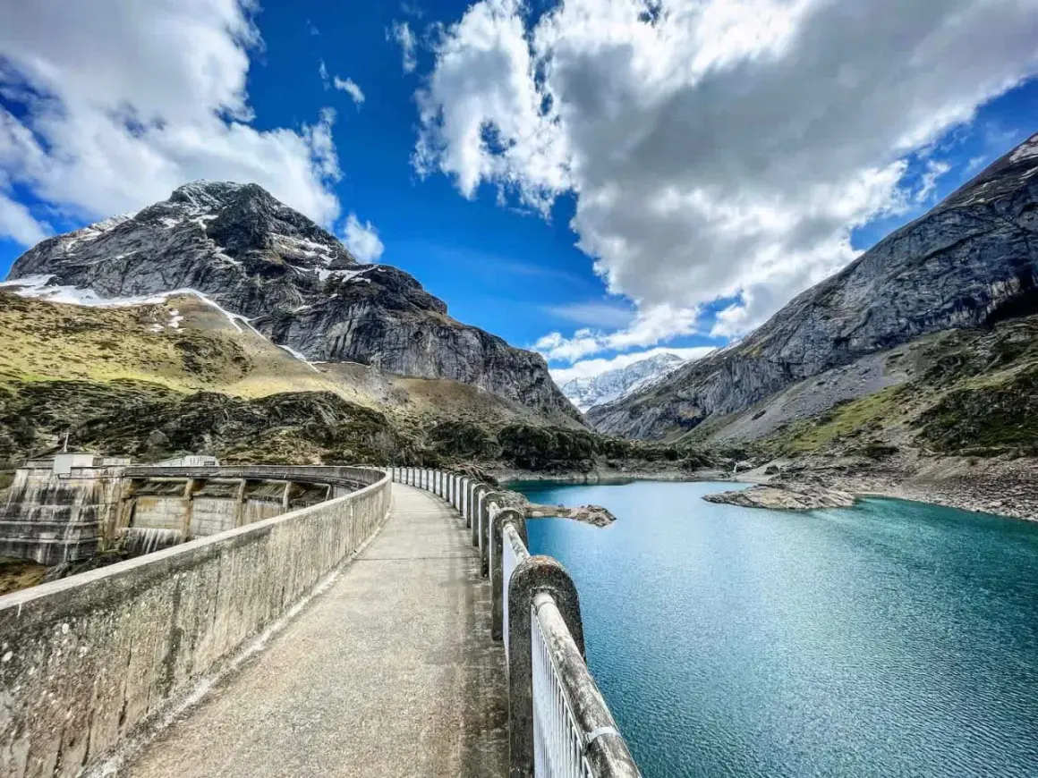 Vue panoramique du lac des gloriettes, un barrage imposant et des montagnes majestueuses sous un ciel bleu et nuageux.