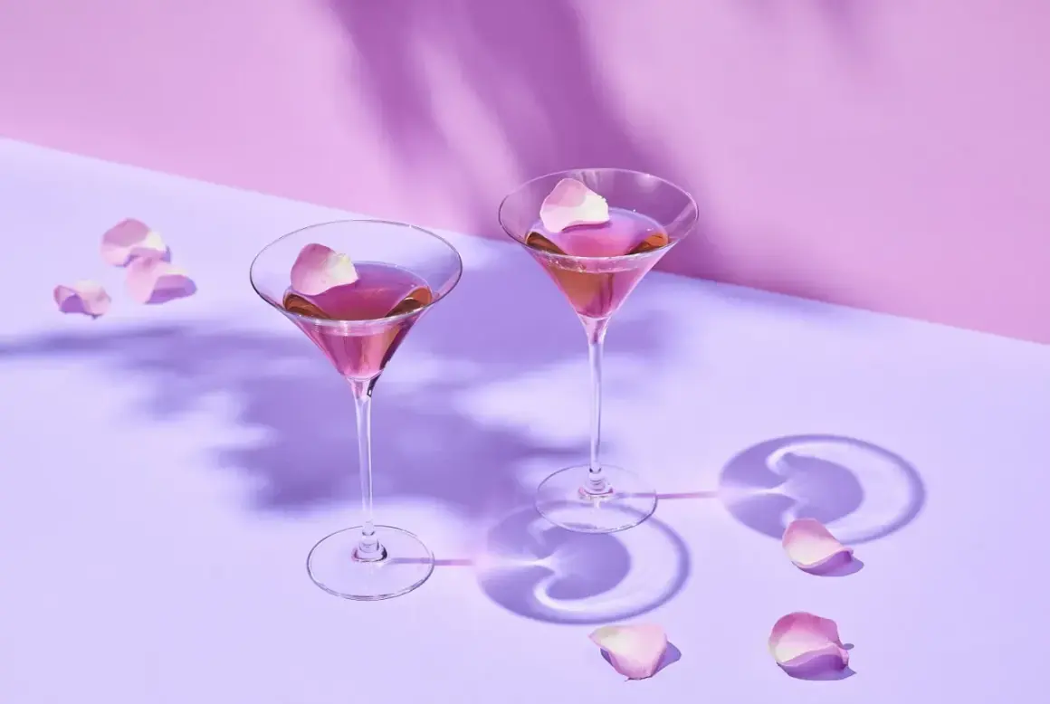 Dwa eleganckie drinki z martini rosato, ozdobione płatkami róż, unoszą się w delikatnym fioletowym świetle.