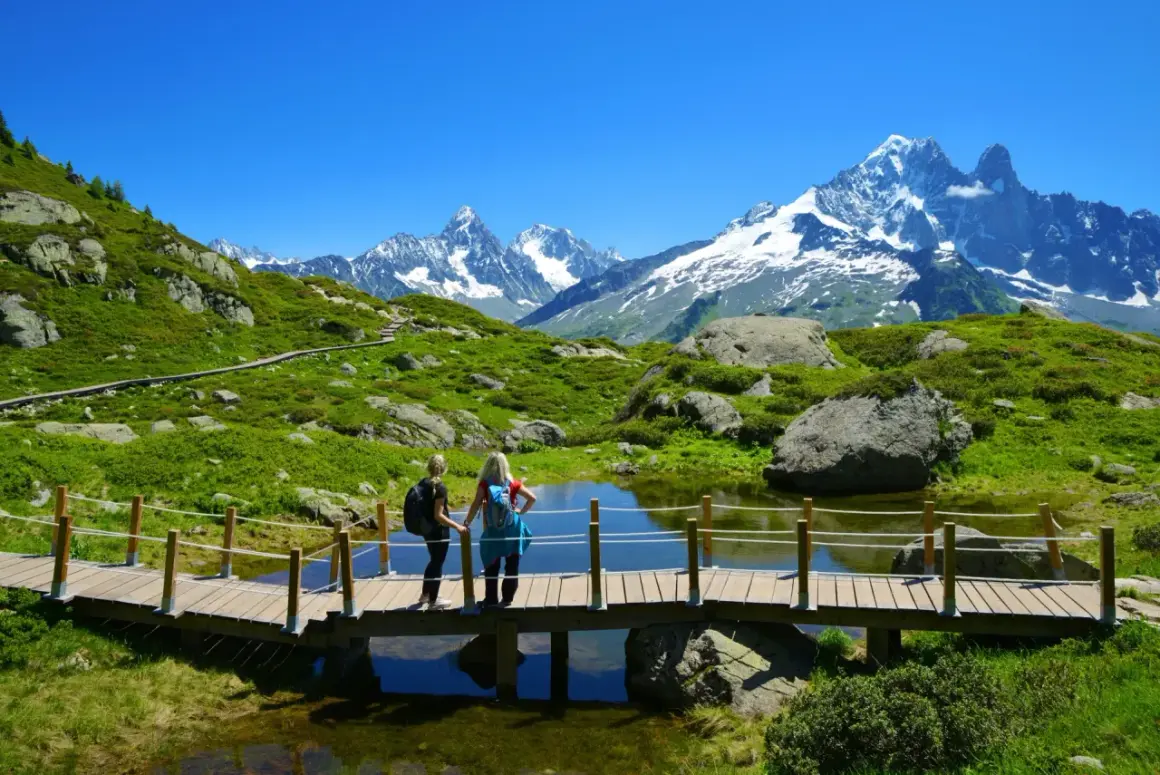 Zwei Wanderinnen auf einer Holzbrücke mit Blick auf den schneebedeckten Mont Blanc und eine grüne Berglandschaft.