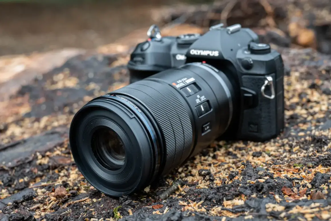 An Olympus camera with a Micro Four Thirds pancake lens rests on a bed of wood chips and dark soil, ready for an outdoor adventure.