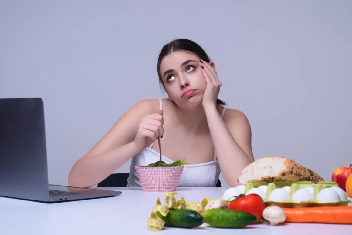 Une jeune femme s'ennuie en mangeant une salade, entourée de nourriture saine. Elle semble regretter de manger trop de fibres.