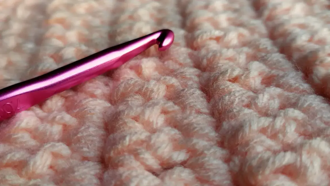 Uncinetto rosa su tessuto lavorato a maglia color pesca, mostrando la differenza tra uncinetto e ferri.