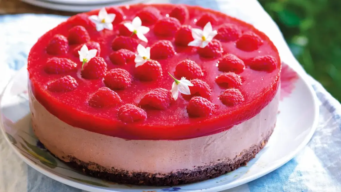 Cheesecake ricetta semplice con lamponi freschi e gelatina rossa, decorata con piccoli fiori bianchi.