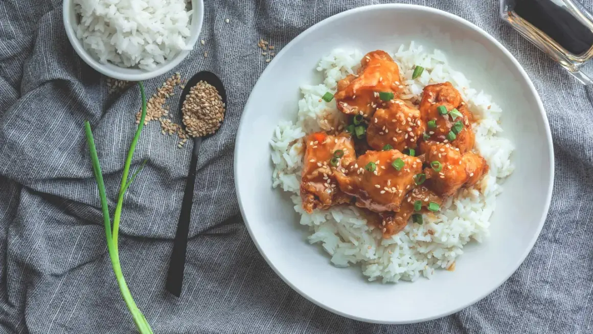 Ein Teller mit Reis und Hähnchen in süß-saurer Sauce, bestreut mit Sesam und Frühlingszwiebeln. Dieses Gericht hat Reis, Proteine und ist ein Genuss.