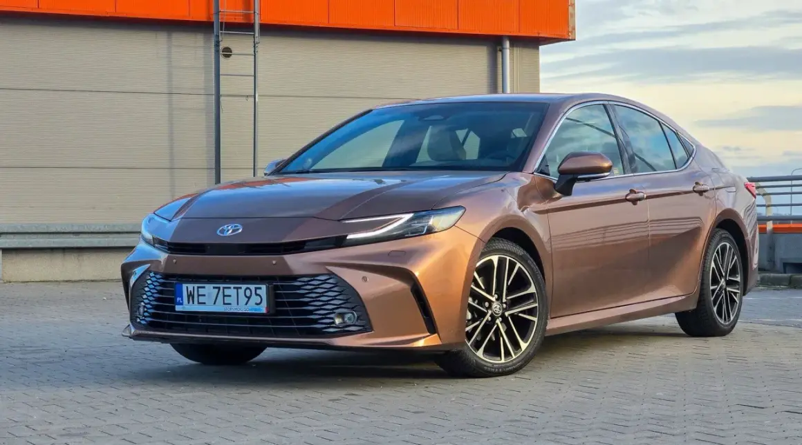 Brązowa Toyota Camry 2.5 Hybrid prezentuje swoje dane techniczne na tle industrialnego budynku.
