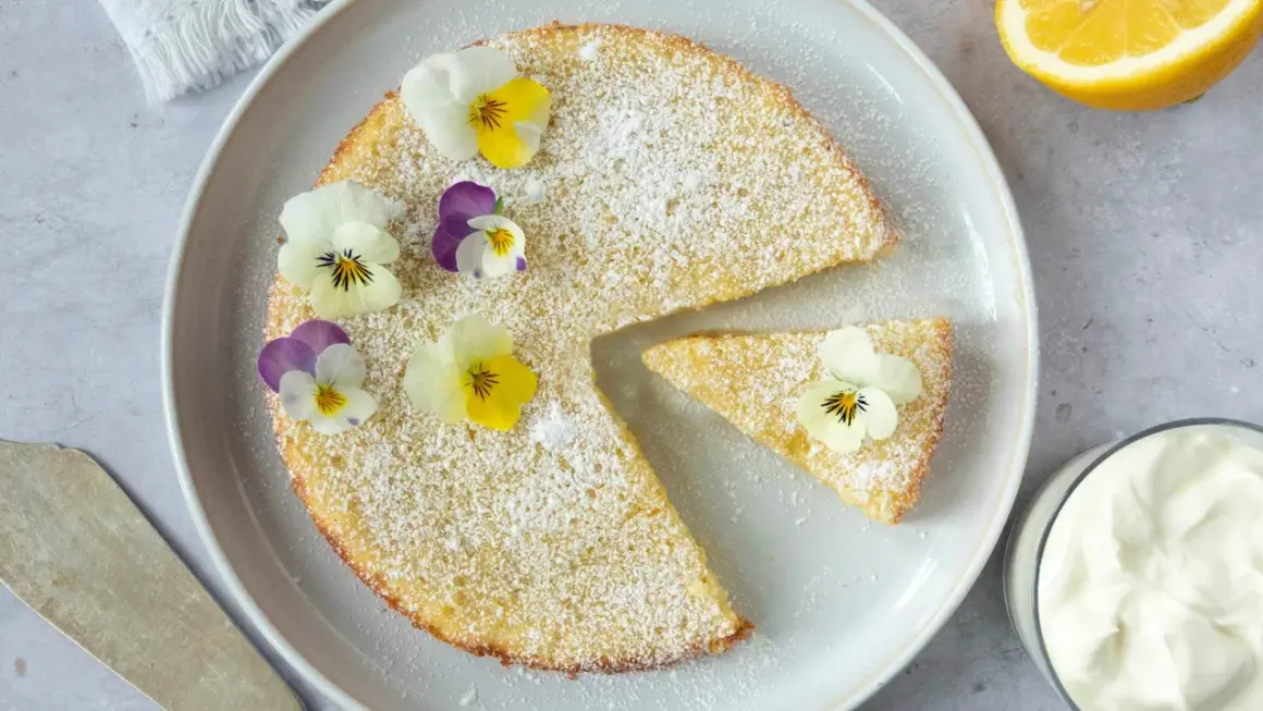En saftig citron kladdkaka pudrad med florsocker och dekorerad med violer. En bit är uppskuren och en skål med grädde syns bredvid.