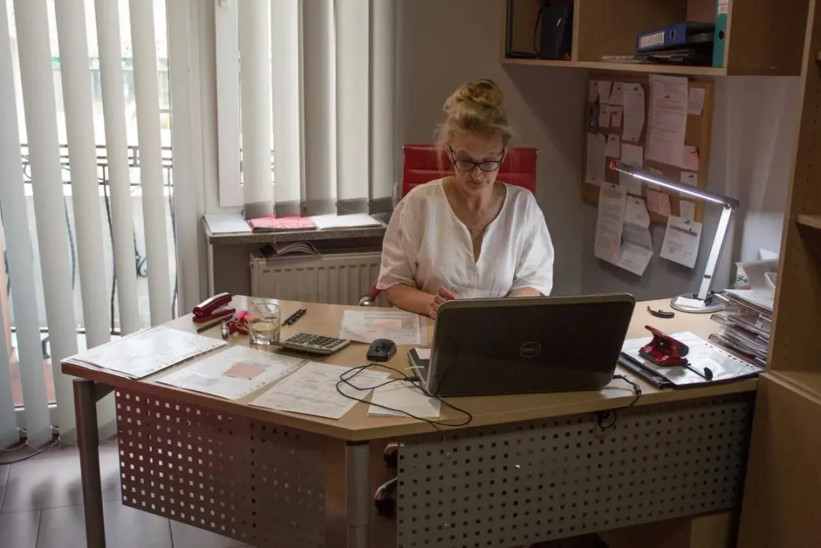 Kobieta w okularach pracuje przy biurku z laptopem, dokumentami i kalkulatorem.