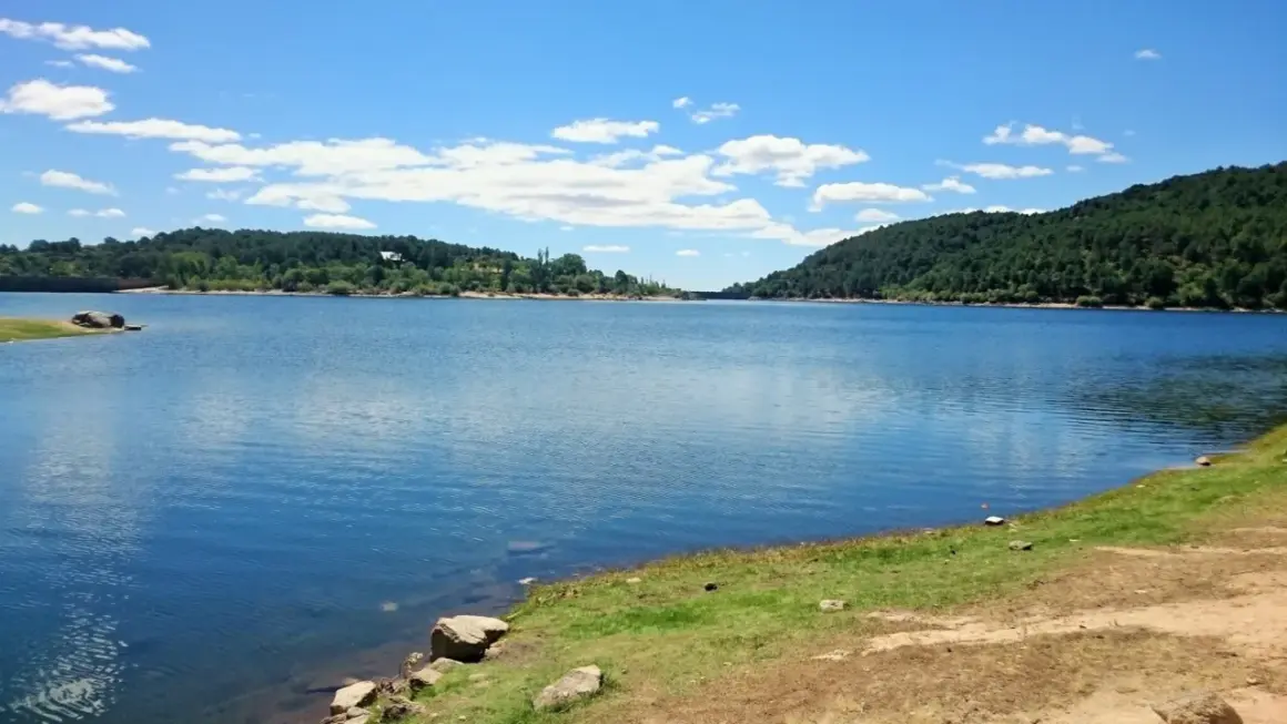 Un día soleado en la ruta del agua La Jarosa, con un embalse azul rodeado de colinas verdes y un cielo con nubes blancas.