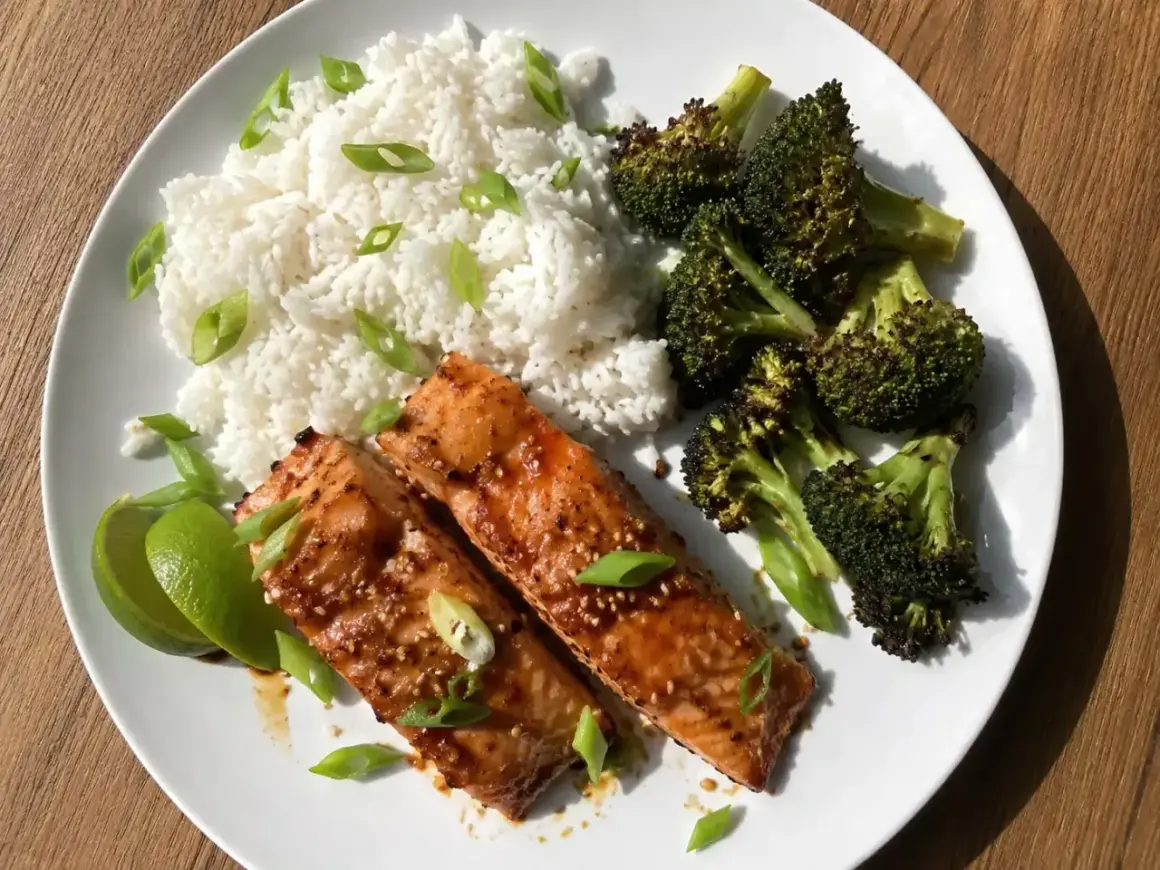 Två filéer av asiatisk lax med sesamfrön, serveras med ris, rostad broccoli och limeklyftor.