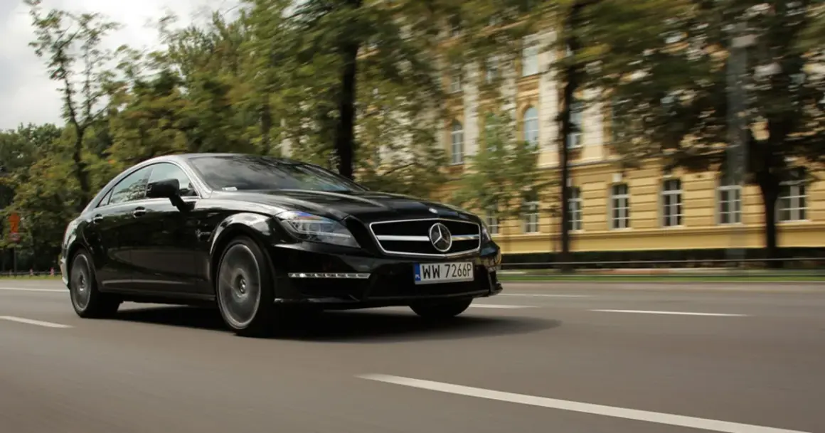 Czarny Mercedes CLS 63 AMG Coupe pędzi ulicą, mijając zabytkową kamienicę i drzewa.