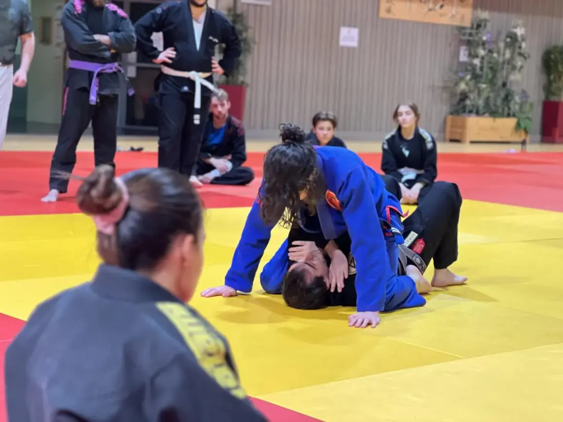 Deux pratiquants de jiu jitsu s'entraînent sur un tapis jaune et rouge. L'un est en position dominante, l'autre en défense.