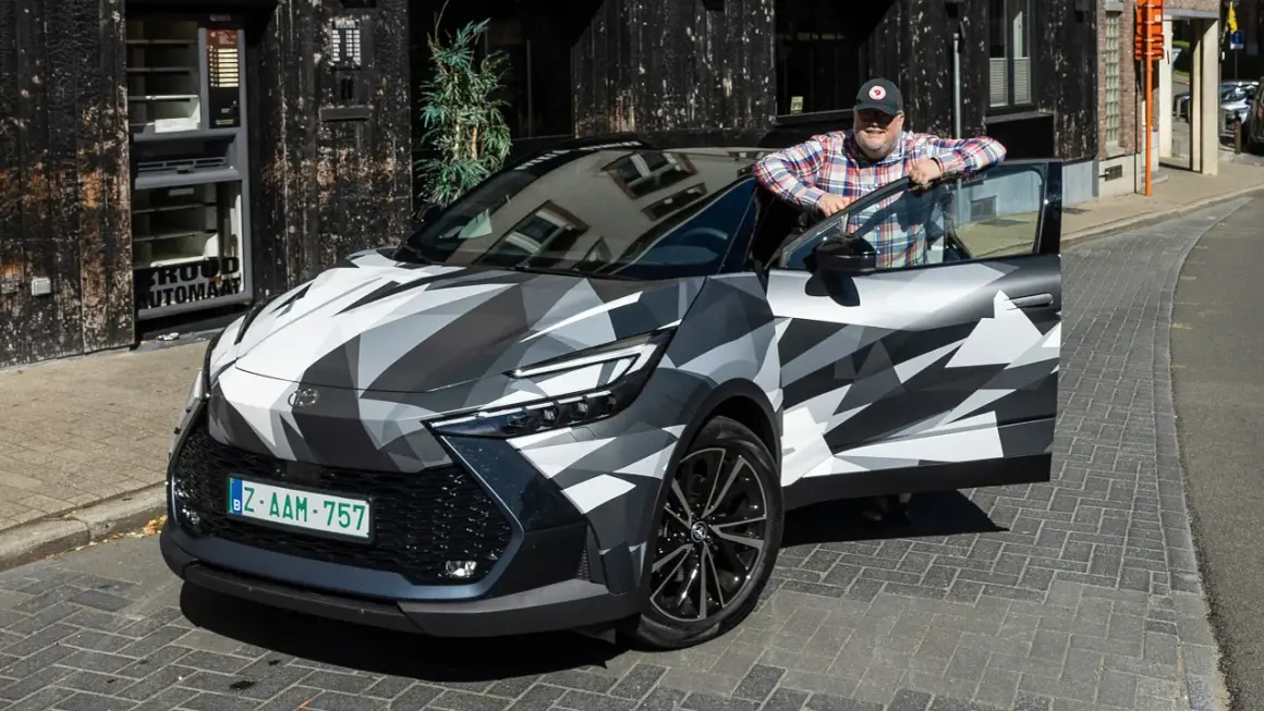 Man lutar sig mot en Toyota C-HR Hybrid i kamouflagemönster. Bilen är parkerad på en kullerstensgata framför en byggnad med en automat.
