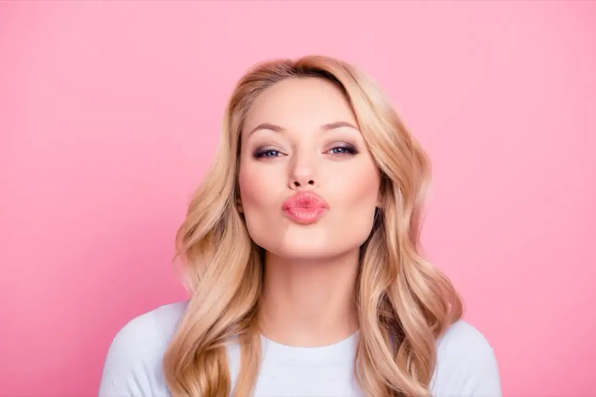 Donna con capelli biondi e trucco labbra naturale, che manda un bacio su sfondo rosa.