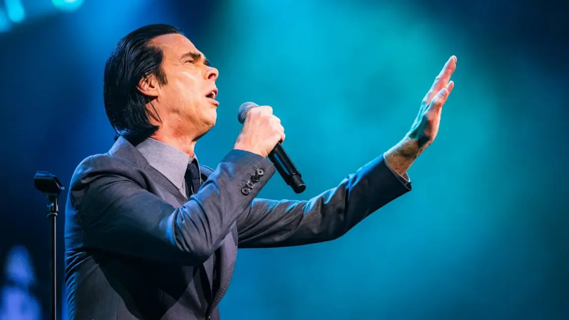 Nick Cave sjunger passionerat med mikrofonen i handen under en nick cave tour. Hans arm är utsträckt mot en blå bakgrund.