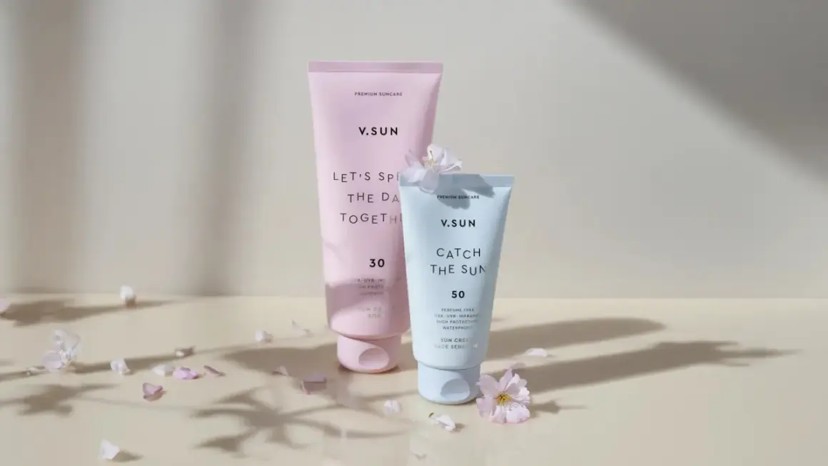 Zwei V.SUN Sonnencreme-Tuben, eine mit LSF 30 für den Körper, die andere mit LSF 50 für das Gesicht, umgeben von zarten Blütenblättern.