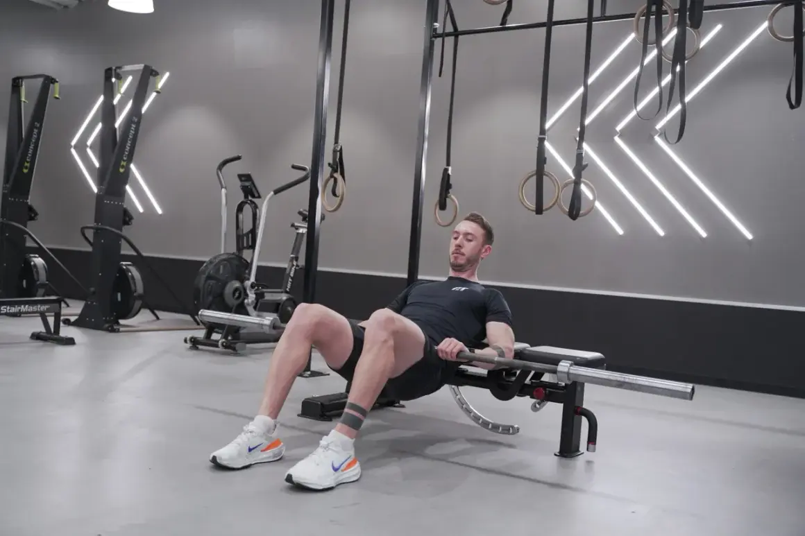 Man utför hip thrusters med skivstång på en bänk i ett modernt gym.
