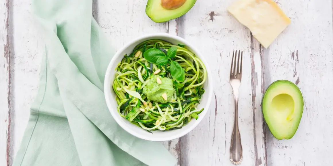 Zoodles con pesto di avocado, guarniti con pinoli e basilico. Accanto, mezzo avocado, un pezzo di parmigiano e una forchetta.