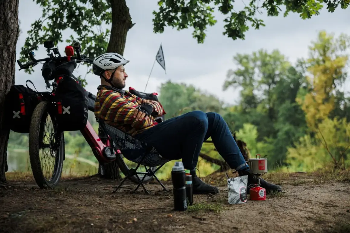 Mężczyzna odpoczywa podczas wycieczki rowerowej, gotując posiłek na kuchence turystycznej. Obok rower z sakwami.