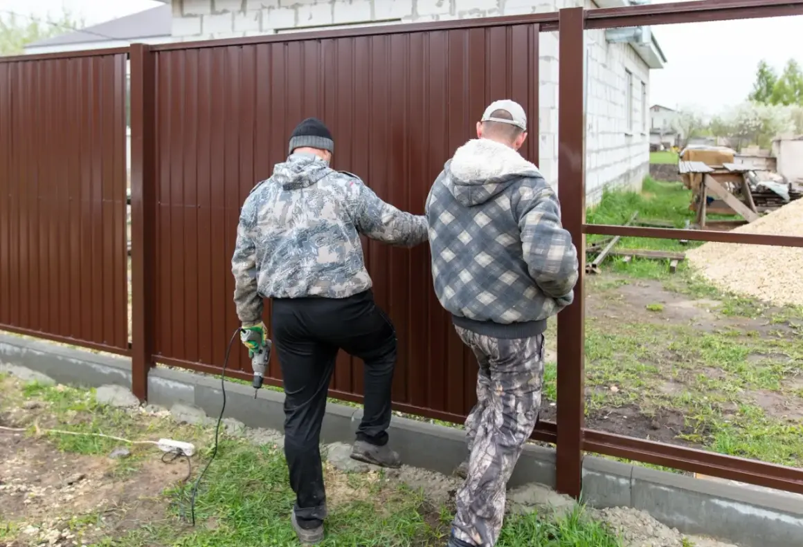 Dwóch mężczyzn montuje brązowe ogrodzenie działki. Sprawdzają zgodność z przepisami budowlanymi.