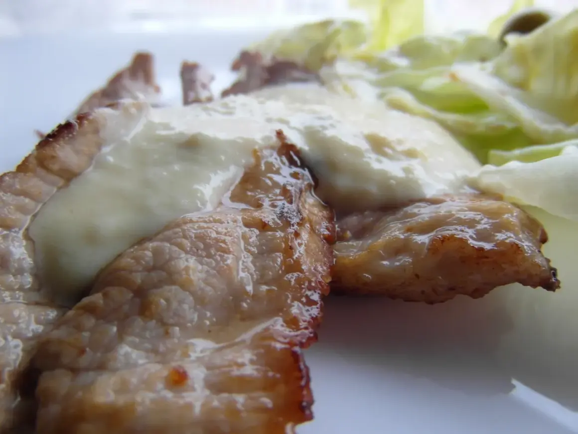 Tiernas lonchas de blanqueta de cerdo cubiertas con una cremosa salsa blanca, acompañadas de hojas verdes frescas.