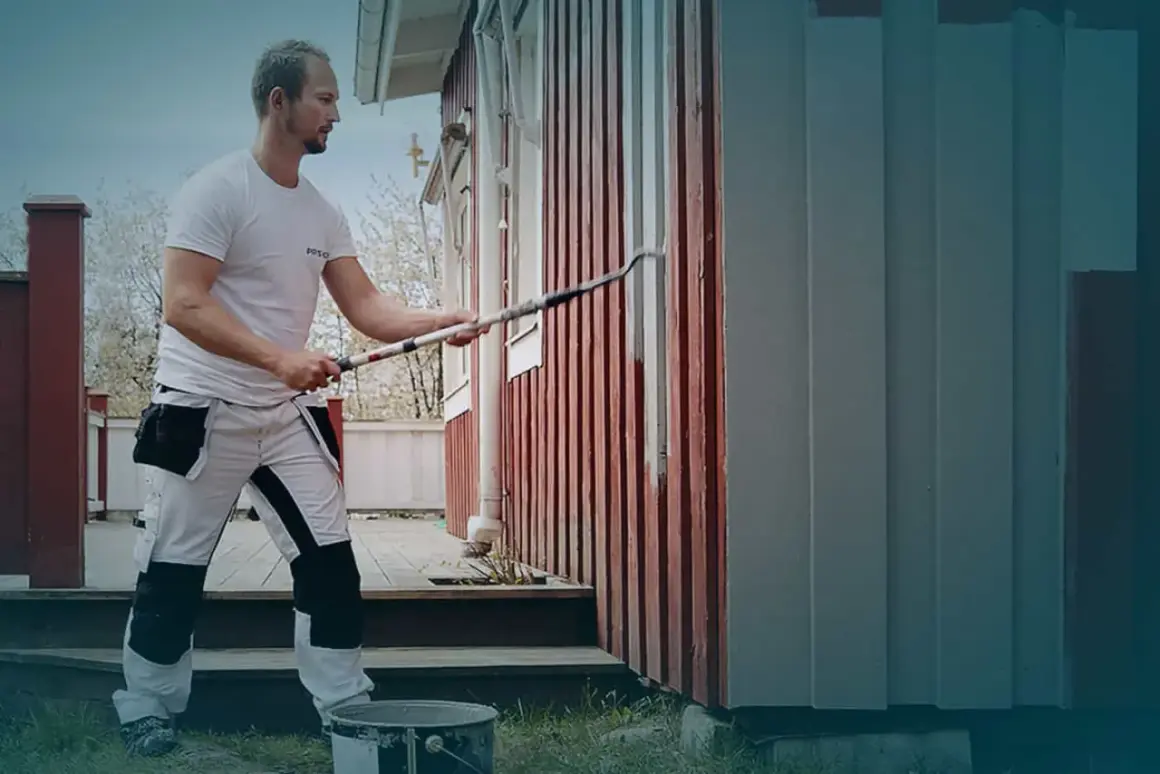 Man målar om huset med en lång roller. Det röda trähuset får nya gråa paneler.