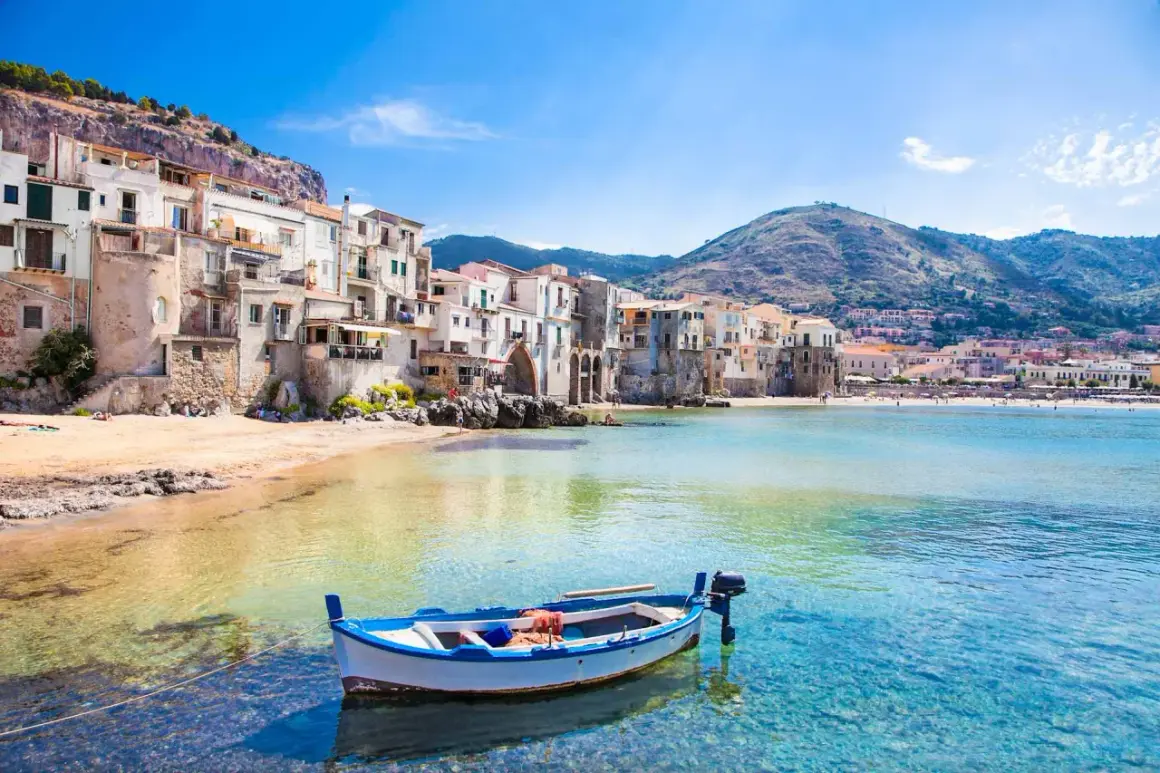 La **spiaggia più bella** di Cefalù, con case colorate a picco sul mare, una barca bianca e blu e acque cristalline.