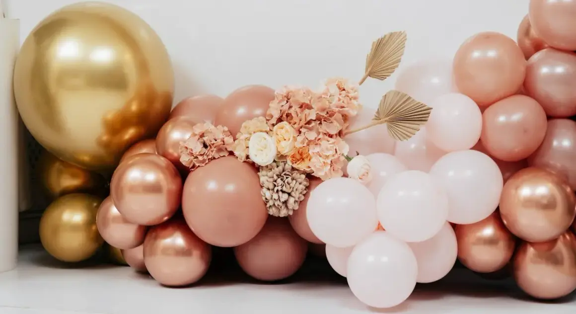 Łuk z balonów w kolorach złota, pudrowego różu i bieli, ozdobiony kwiatami.
