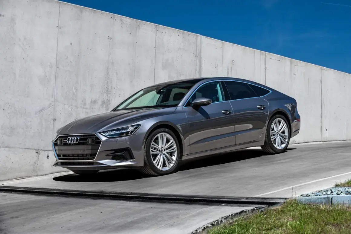 Elegancki, szary Audi A7 2025 z panoramicznym dachem stoi na tle betonowej ściany.