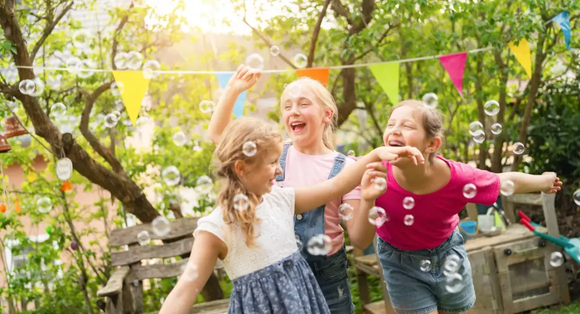 Trzy dziewczynki śmieją się i łapią bańki mydlane w ogrodzie. Wesołe życzenia urodzinowe dla dziecka!